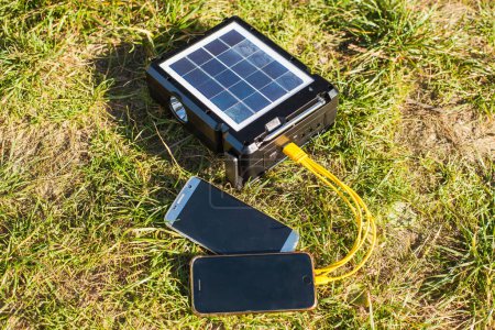 Portable power bank with a solar panel for recharging gadgets while camping. The solar panel lies on the green grass under the sun and charges two phones at once.