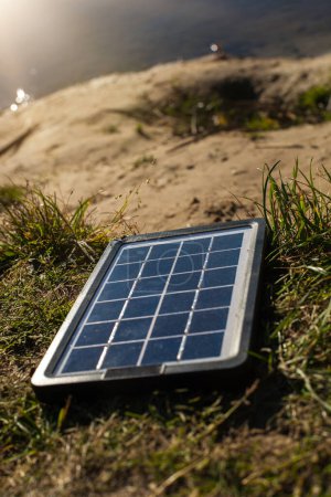 Téléchargez les photos : Photo verticale d'un panneau solaire dans la nature. La station se trouve sur le fond d'herbe, de sable et d'eau. Les rayons du soleil pénètrent dans le cadre et inondent le panneau solaire de son éclat - en image libre de droit