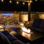 A resort style backyard at night with a waterfall, pergola, and a firepit at night.