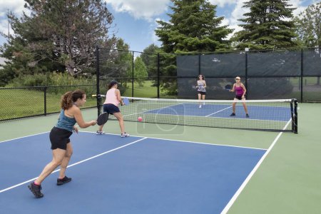 A comptetivie doubles game of pickleball with a group of women on a blue and green court in summer.