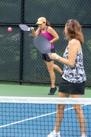A pickleball player returns a serve as her partner looks on at a suburban pickleball court during summer.