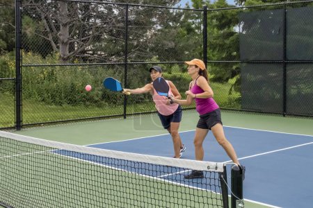 A comptetivie doubles game of pickleball with a group of women on a blue and green court in summer.