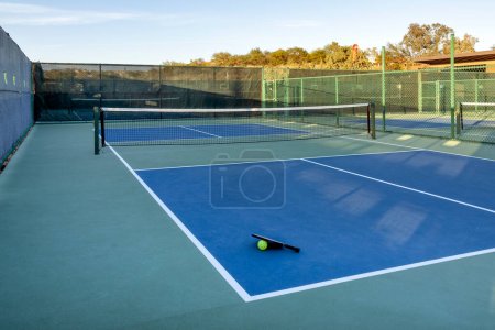 A pickleball paddle and yellow ball are shown on an empty court at the end of an active day.