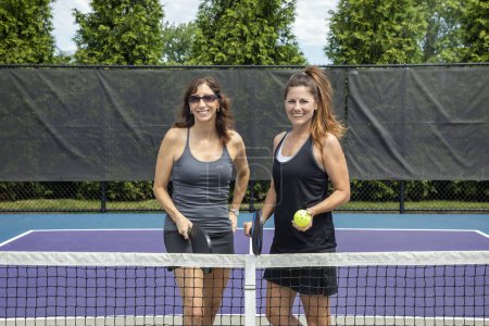 Two pickleball players posing with paddles and a ball in front of the net on a court during summer.