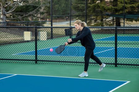 A female pickleball player prepares to serve a bright pink ball on a blue and green court in early spring.