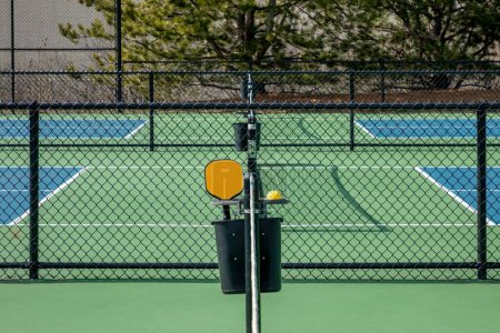 View of an orange pickleball paddle and yellow ball in between blue and green courts with black fencing at a suburban complex.
