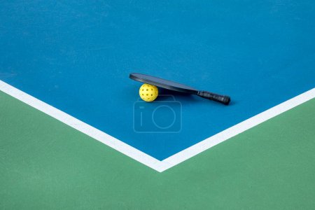 View of a pickleball paddle and yellow ball on a blue and green court with white lines.