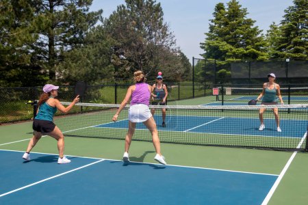 A comptetivie doubles game of pickleball at the net on a blue and green court in summer.