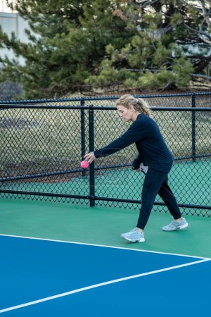 A female pickleball player prepares to serve a bright pink ball on a blue and green court in early spring.