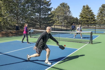 A player hits a backhand in a comptetivie doubles game of pickleball with a group of men and women on a blue and green court in spring.