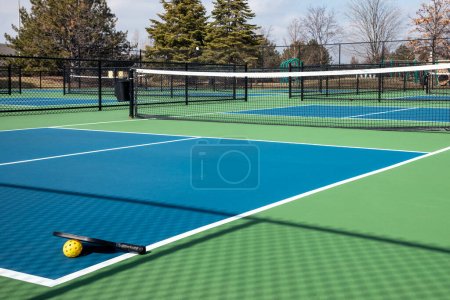 View of a pickleball complex with a paddle and yellow on blue and green courts beside a playground in a suburban park in early spring.