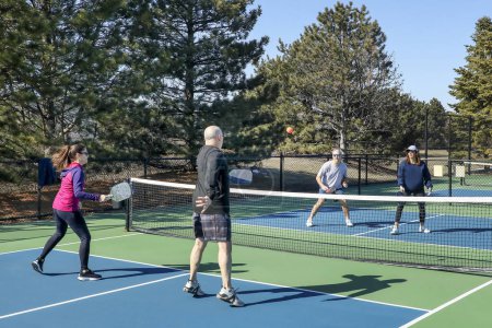 A comptetivie doubles game of pickleball at the net with a group of men and women on a blue and green court in spring.