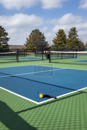 View of a pickleball complex with a paddle and yellow ball on blue and green courts beside a playground in a suburban park in early spring.