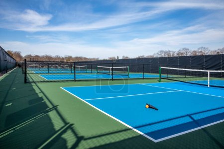 View of a new pickleball complex with a paddle and orange ball on blue and green courts in a suburban setting during early spring.