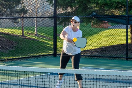 A female pickleball player returns a volley of a bright yellow ball at the net on a dedicated court at a public park.