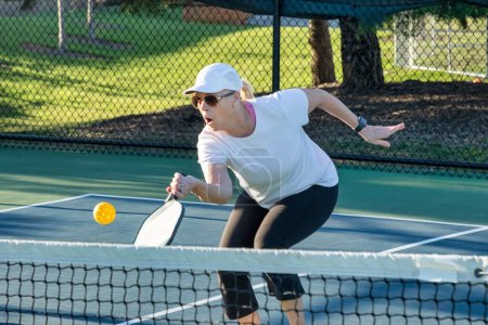 A female pickleball player returns a volley of a bright yellow ball at the net on a dedicated court at a public park.