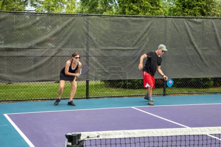 A pickleball player serves while his partner prepares for a return on a suburban pickleball court during summer.