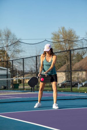 A female pickleball player serves a bright pink ball at the baseline on a dedicated court at a public park.