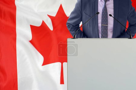 Tribune with microphone and man in suit on Canada flag background. Businessman and tribune on Canada flag background. Politician at the podium with microphones background Canada flag. Conference