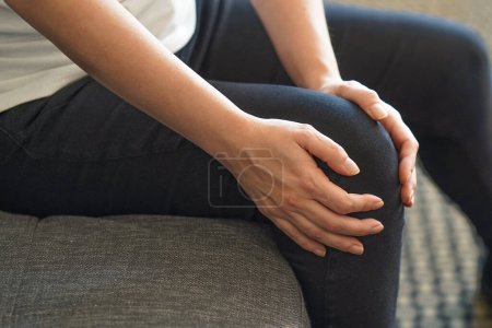 Foto de Mujer de cerca sentada en el sofá y sintiendo dolor de rodilla y se masajea la rodilla en casa. Salud y concepto médico - Imagen libre de derechos