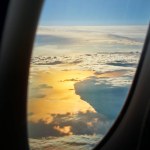 Sunset photo above the clouds through the oval airplane window, wonderful play of colors in the golden orange evening light over the sea, the fluffy clouds and the land, square