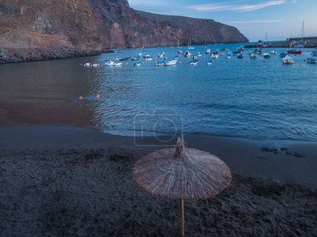 Téléchargez les photos : Coucher de soleil sur la plage de sable noir Playa de Vueltas avec parasol, bateaux de pêche et falaises d'ouk. Valle Gran Rey, La Gomera, Îles Canaries, Espagne. - en image libre de droit