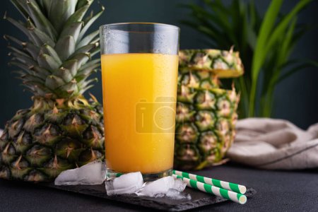 Pineapple juice in a glass with ice and fresh pineapple pieces on the dark background