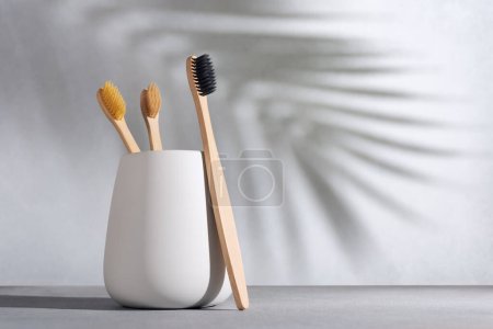 Téléchargez les photos : Brosse à dents écologique naturelle avec manche en bambou en bois dans une tasse blanche sur un fond avec ombre de palmier. Style de vie zéro déchet. - en image libre de droit