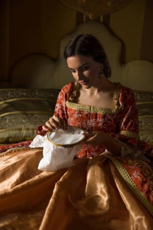 Téléchargez les photos : Chambre médiévale dorée avec beau lit à baldaquin. Une dame en robe renaissance faisant de la broderie. - en image libre de droit