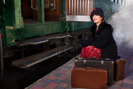 Photo for Attractive young woman in red 1920s flapper dress and cloche hat travelling by antique steam train - Royalty Free Image