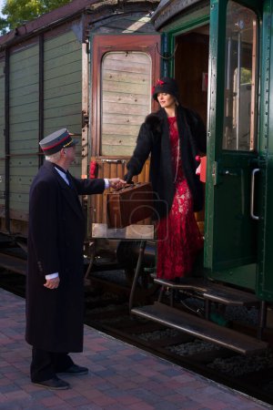 Photo for Attractive young woman in red 1920s flapper dress and cloche hat travelling by antique steam train - Royalty Free Image