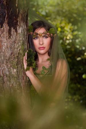 Belle jeune femme en robe verte se faisant passer pour une fée de la forêt avec des tissus en mousseline de soie pure et des guirlandes de mousse