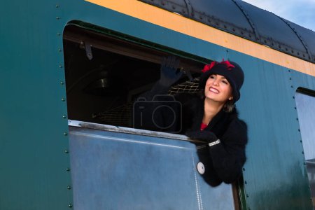 Photo for 1920s woman wearing fur coat and cloche hat hanging out of the window of an antique train carriage - Royalty Free Image