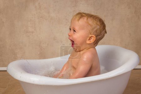 Téléchargez les photos : Scènes heureuses d'un petit garçon mignon de 12 mois assis dans une baignoire blanche vintage éclaboussant d'eau et de savon - en image libre de droit