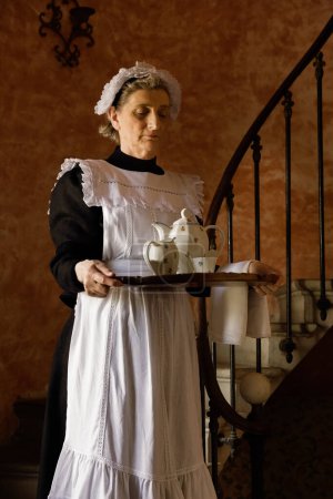 Criada victoriana o sirvienta en vestido negro, gorra de encaje y delantal blanco trabajando en un interior del siglo XIX