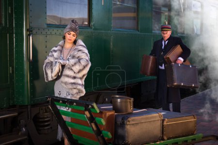 Téléchargez les photos : Scène de reconstitution sur le quai près d'un authentique wagon de train de première classe 1927 où une dame des années 1920 chic attend ses bagages - en image libre de droit