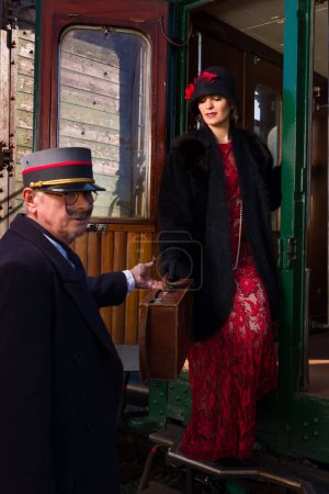 Téléchargez les photos : Attractive young woman in red 1920s flapper dress and cloche hat travelling by antique steam train - en image libre de droit