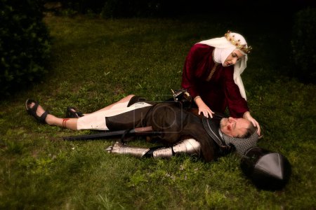 Escena medieval de una joven reina viuda llorando arrodillada junto a un caballero muerto caído en el campo de batalla