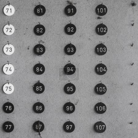 Schwarz-Weiß-Aufnahme einer Reihe von runden Zahlenanhängern, die in Reihen und Spalten an einer Wand angeordnet sind.
