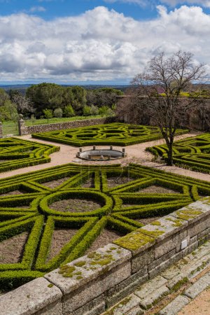 The Royal El Escorial Monastery's gardens display precise geometric hedges against a scenic backdrop, under the dramatic Madrid sky