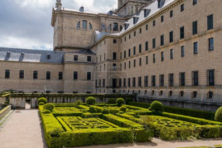 The Royal El Escorial Monastery's gardens display precise geometric hedges against a scenic backdrop, under the dramatic Madrid sky