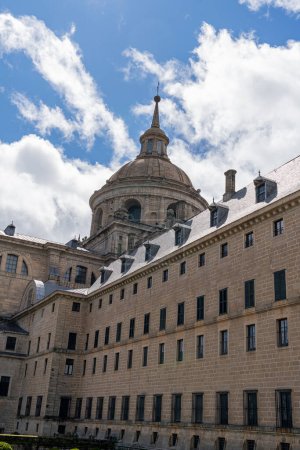 Sculptural details and the iconic dome of El Escorial Monastery stand out against a blue sky with fluffy clouds