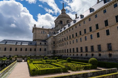 Die Gärten des Königlichen El Escorial-Klosters zeigen präzise geometrische Hecken vor einer malerischen Kulisse unter dem dramatischen Himmel von Madrid