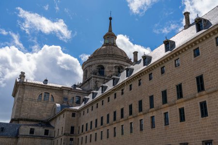 Foto de Los detalles escultóricos y la icónica cúpula del Monasterio de El Escorial destacan sobre un cielo azul con nubes esponjosas - Imagen libre de derechos