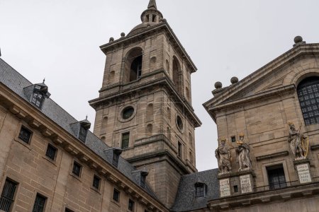 Statuen historischer Persönlichkeiten stehen über dem Haupthof des El Escorial-Klosters, vor der strengen Eleganz der Fassade des Gebäudes.