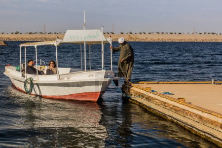 Foto de ASWAN, EGYPT - FEB 13, 2019: Boat near Kalabsha temple in the Nasser lake with the Aswan high dam in the background, Egypt - Imagen libre de derechos