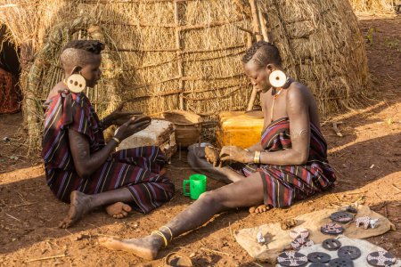 Foto de OMO VALLEY, ETIOPÍA - 6 DE FEBRERO DE 2020: Muchachas de la tribu Mursi preparan placas labiales en su aldea, Etiopía - Imagen libre de derechos