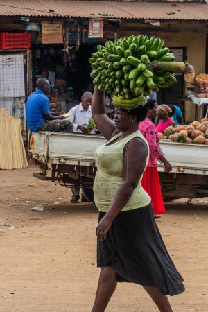 Foto de BUDADIRI, UGANDA - 25 DE FEBRERO DE 2020: Mujer llevando racimo de plátanos matoke en la aldea de Budadiri, Uganda - Imagen libre de derechos