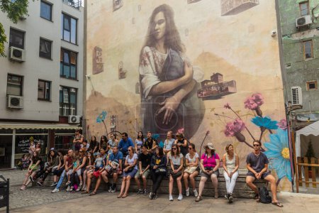 Foto de PLOVDIV, BULGARIA - 29 de julio de 2019: La gente se sienta debajo de un graffiti en una pared en el distrito creativo de Kapana en Plovdiv, Bulgaria - Imagen libre de derechos
