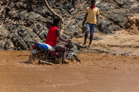 Foto de OMO VALLEY, ETIOPÍA - 4 DE FEBRERO DE 2020: Niño local en una motocicleta cruzando aguas hinchadas del río Kizo, Etiopía - Imagen libre de derechos